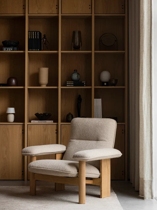 Audo Copenhagen Brasilia Lounge Chair Natural Oak Moss in a cozy living room setting with wooden shelving.